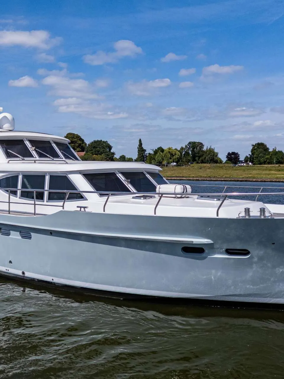 Een witte en lichtgrijze motorboot vaart op een waterweg met een groene oever en blauwe lucht op de achtergrond.