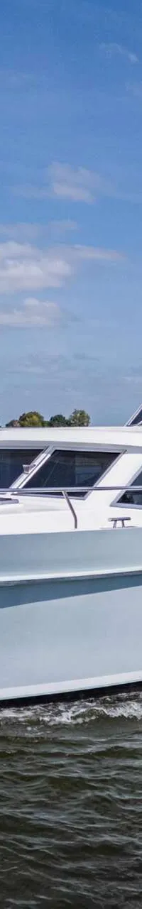 Een witte motorjacht met een lichtblauwe romp vaart op een waterweg onder een blauwe hemel met wolken.