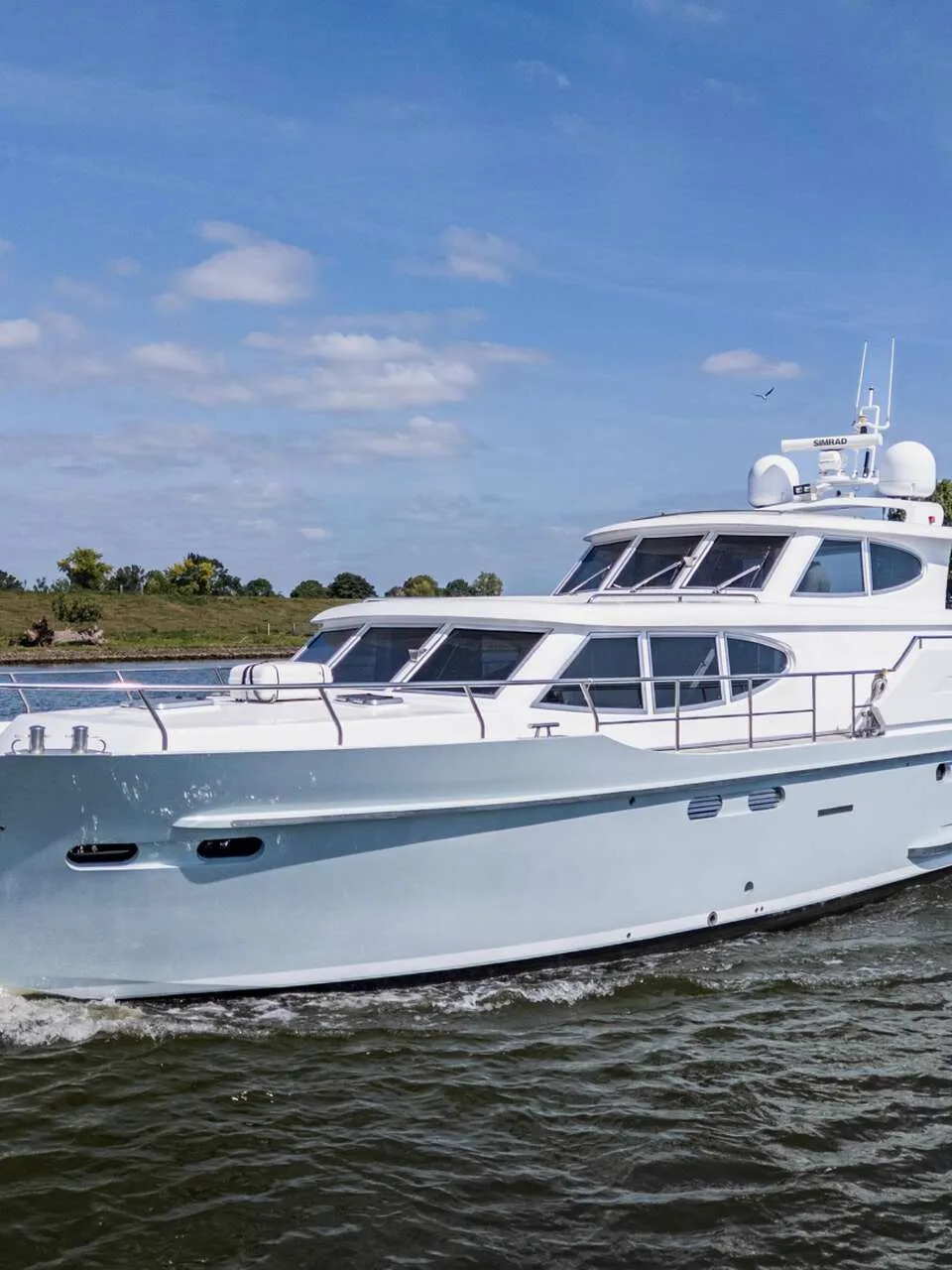 Een witte motorjacht met een lichtblauwe romp vaart op een waterweg onder een blauwe hemel met wolken.