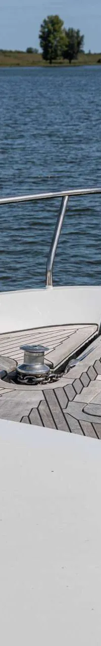 De foto toont het voordek van een boot met teakhouten dek, een ankerlier, reling, bolders en een luik.