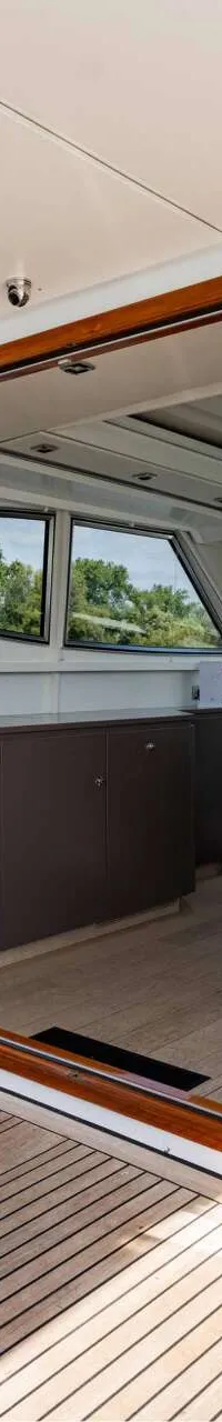 Een houten tafel met twee rieten stoelen op een teakhouten dek, met uitzicht op water en bomen, en een glazen schuifdeur…