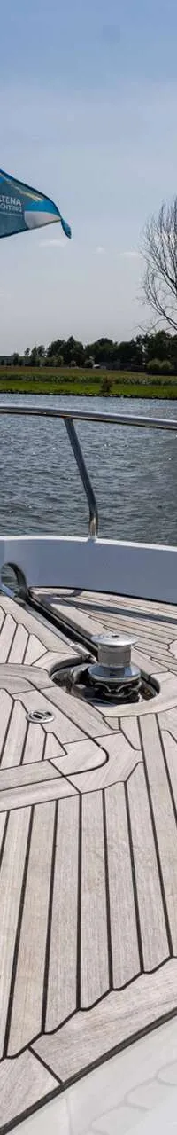 De foto toont de boeg van een boot met een houten dek, reling, twee bolders en een vlaggenmast met een blauwe vlag.