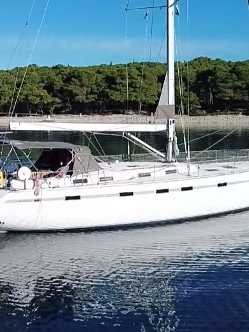 Een witte zeilboot met een grijze kap ligt op rustig water, met een beboste kustlijn op de achtergrond. — Bavaria 55 Cruiser