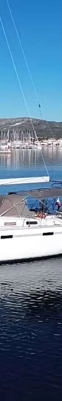 Een witte zeilboot met de naam "ALDEBARAN" ligt op rustig water, met een jachthaven en een stadje op de achtergrond.