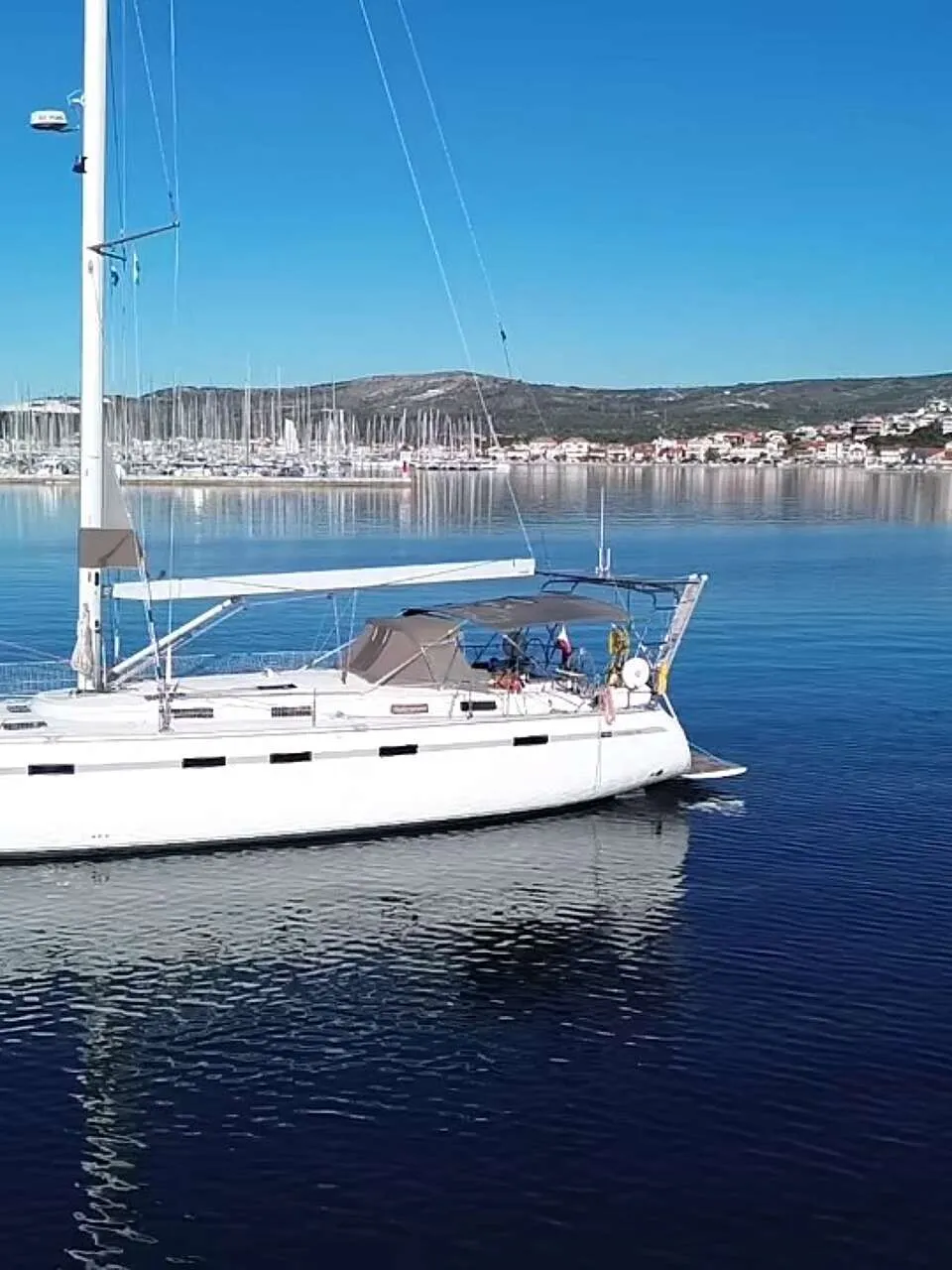 Een witte zeilboot met de naam "ALDEBARAN" ligt op rustig water, met een jachthaven en een stadje op de achtergrond.
