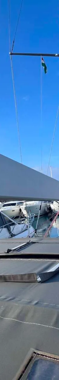 Een zeilboot met een grijs dekzeil en een witte giek met "Seldén" erop, ligt in een jachthaven onder een blauwe lucht.