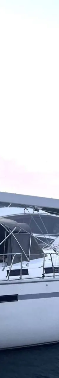 Een witte zeilboot met de tekst "CRUISER 55" ligt aangemeerd in een haven, met andere boten en een heuvelachtige kustlijn…