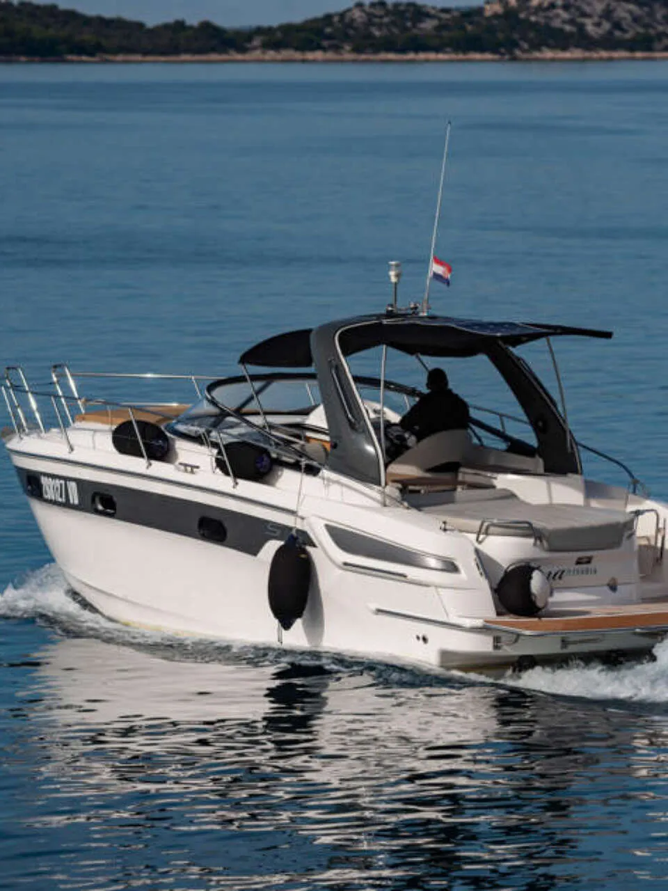 Een witte motorboot met een zwarte streep vaart over blauw water, met een persoon aan het roer en een vlag op het dak.