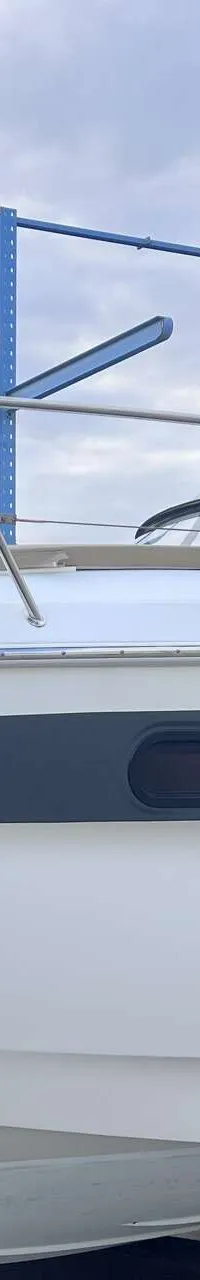Een witte boot met een donkergrijze streep en patrijspoorten staat op een trailer onder een bewolkte hemel. — Bavaria S29