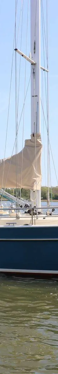 Een blauwe zeilboot met een beige zeil en een beige opbouw vaart op het water, met een persoon aan het roer.