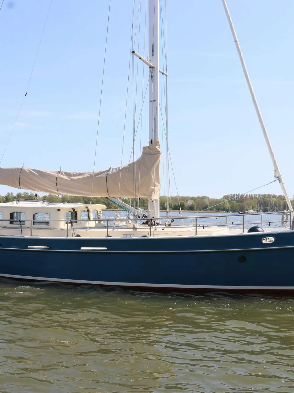 Een blauwe zeilboot met een beige zeil en een beige opbouw vaart op het water, met een persoon aan het roer.