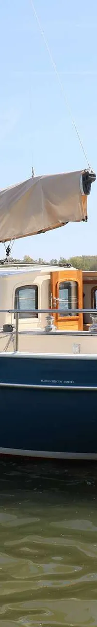 Een blauwe zeilboot met een beige kajuit en een opgerold zeil, met een persoon aan boord en een Nederlandse vlag aan de…