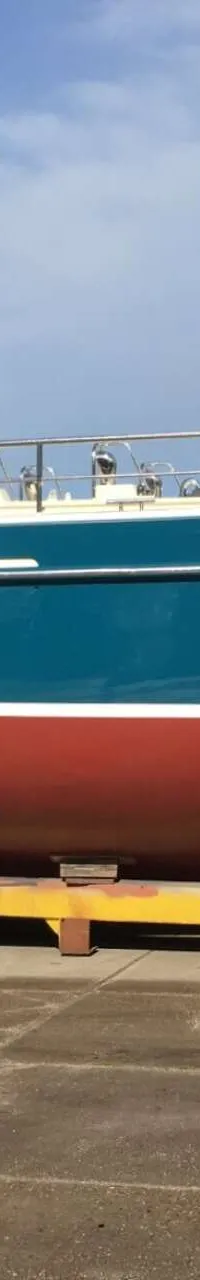Een blauw-witte boot met een rode onderkant staat op een gele trailer op een betonnen ondergrond onder een blauwe lucht.