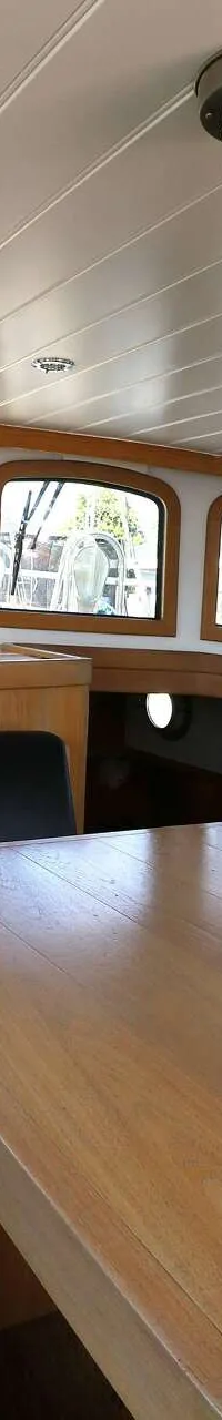 De foto toont een interieur van een boot met een houten tafel, een zwarte leren bank, ramen met gordijnen en een stuurwiel.