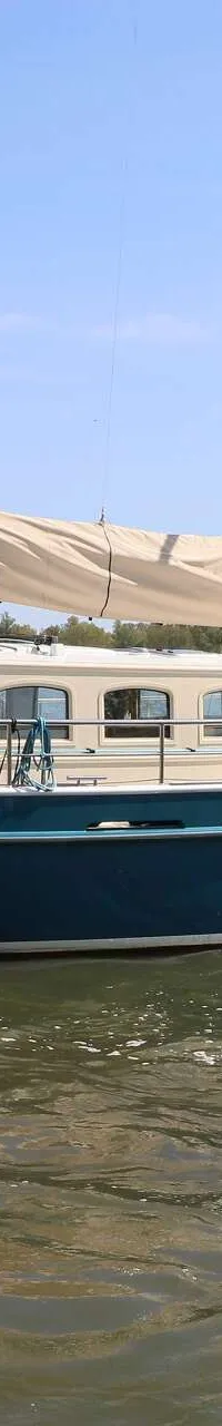 Een blauwe zeilboot met een beige kajuit en opgerold zeil vaart op het water, met een persoon aan het roer.