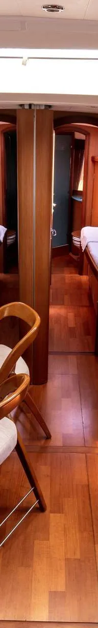 De foto toont het interieur van een boot met een houten vloer, twee banken, een eettafel met servies en twee deuren die…