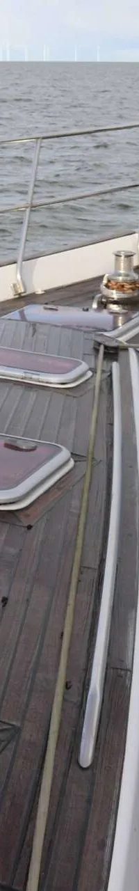 Een houten dek met luiken, een reling en een lier, met de zee en windmolens op de achtergrond.