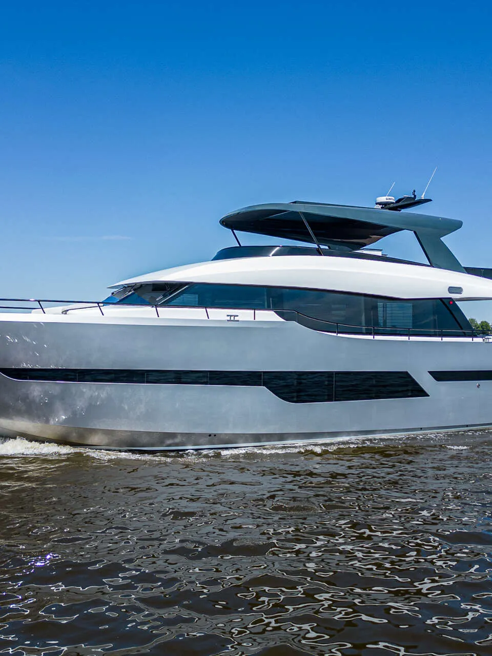 Een grote, grijze motorboot met een witte opbouw vaart op een waterweg onder een helderblauwe lucht.