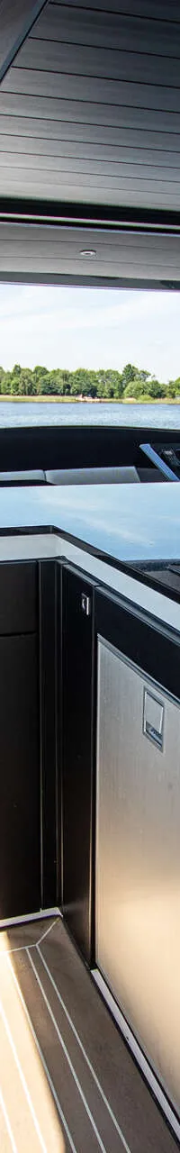 Een bootinterieur met een kombuis, stuurstand en zitgedeelte, met uitzicht op water en een beboste oever.