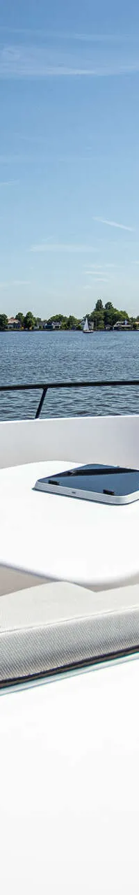 De foto toont het voordek van een boot met zitkussens en een houten dek, varend op water met een dorp op de achtergrond.