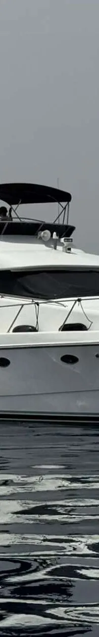 Een witte motorboot met een zwarte bimini en afdekzeilen vaart op rustig water, met een stad en bergen op de achtergrond.