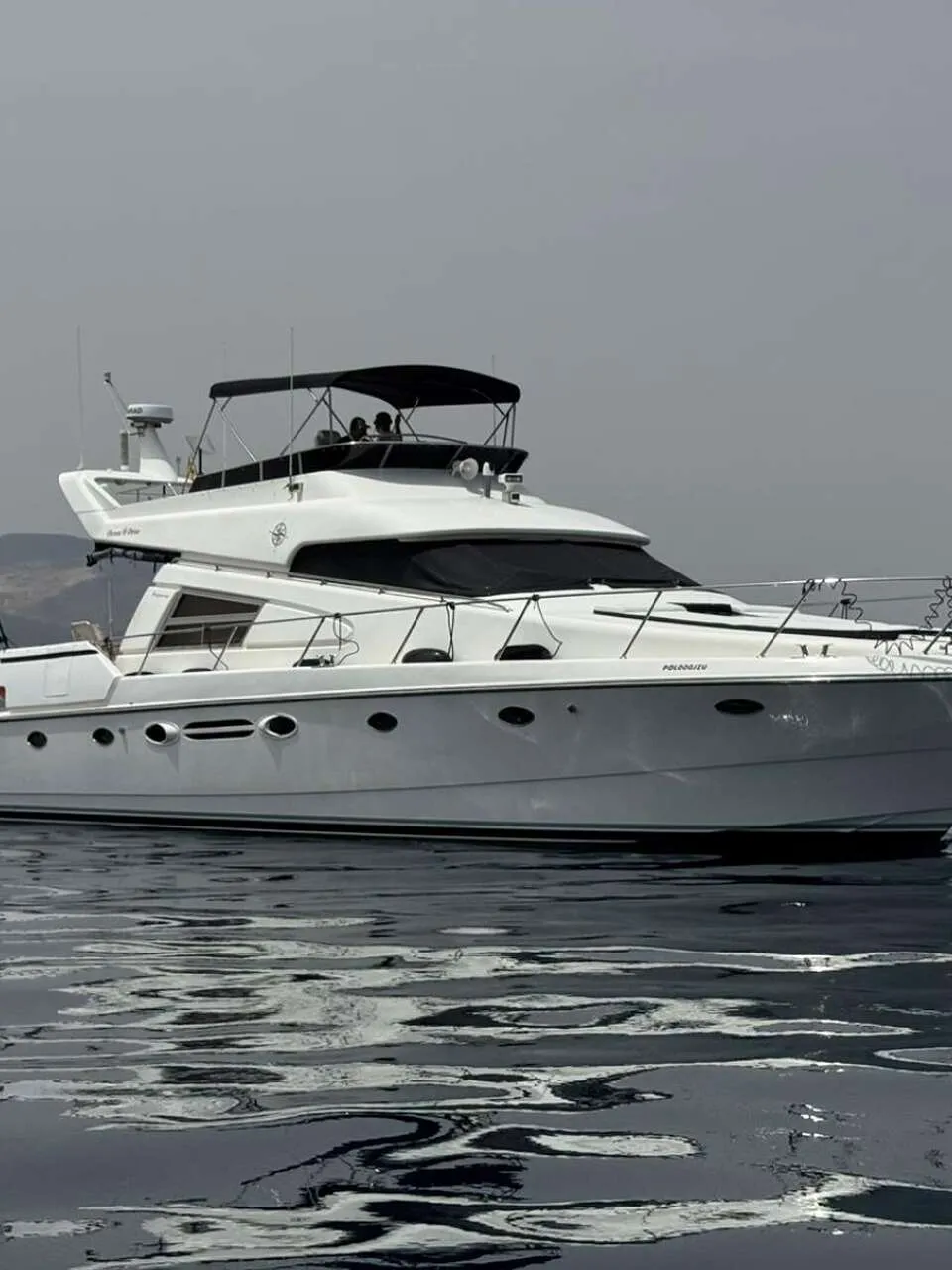 Een witte motorboot met een zwarte bimini en afdekzeilen vaart op rustig water, met een stad en bergen op de achtergrond.