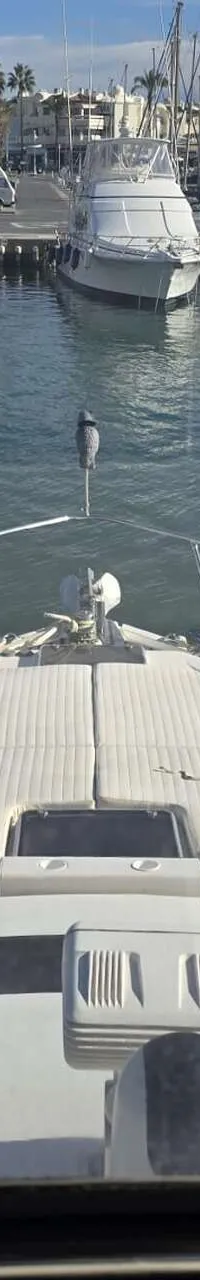 De foto toont het voordek van een witte boot met reling, liggend in een haven met andere boten en gebouwen op de achtergrond.