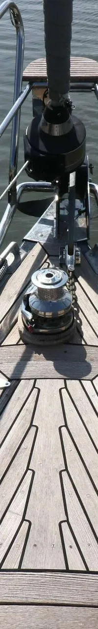 De foto toont de boeg van een zeilboot met een houten dek, reling, ankerlier en touwen, liggend aan een houten steiger.