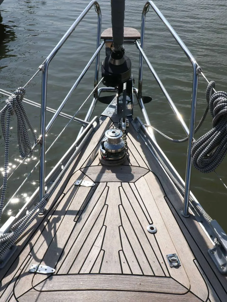De foto toont de boeg van een zeilboot met een houten dek, reling, ankerlier en touwen, liggend aan een houten steiger.