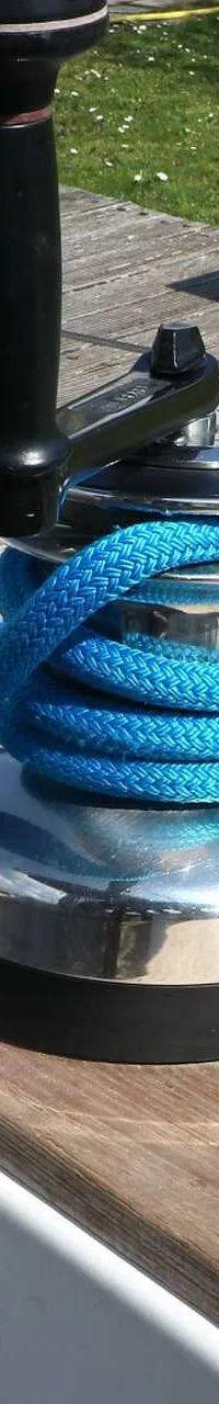 Een glimmende lier met blauw touw en een lierhendel op een houten dek, met een steiger op de achtergrond.
