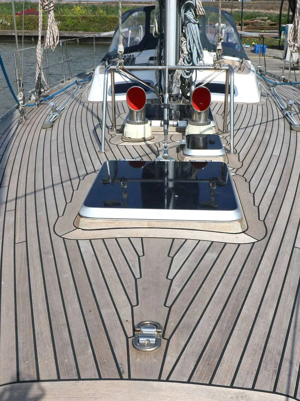 Een teakdek met een luik, twee rode ventilatiekappen en een mast met touwen.