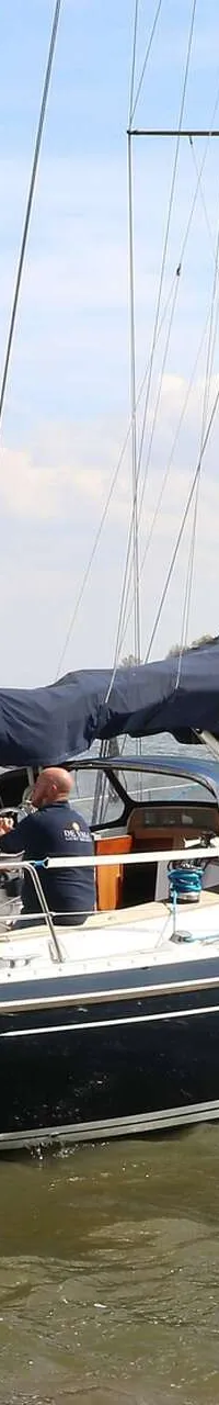 Een donkerblauwe zeilboot met de naam "Indigo" en "Monnickendam" op de spiegel, vaart op water met een Nederlandse vlag aan…