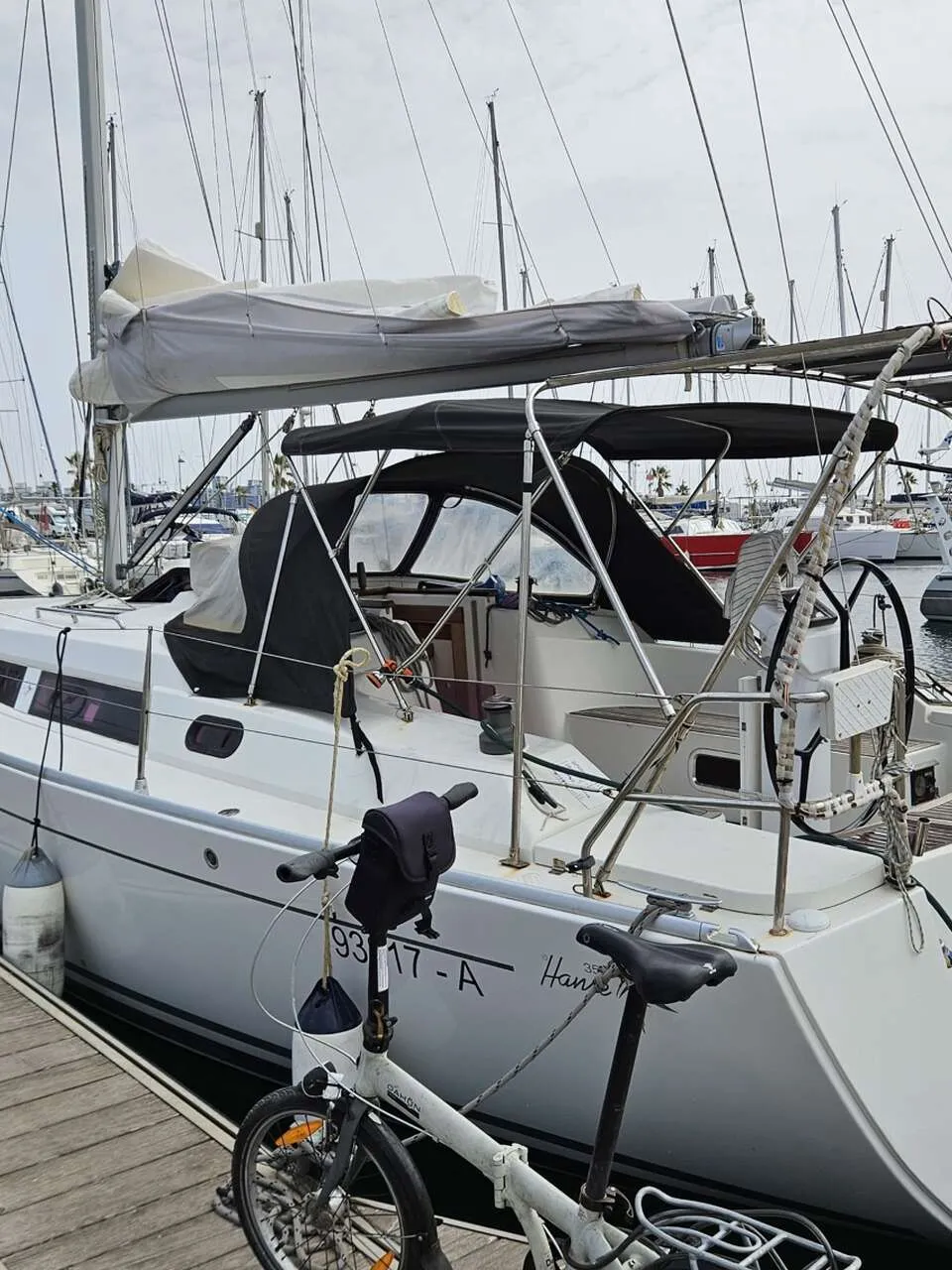 Een witte zeilboot met een zwarte buiskap en bimini ligt aangemeerd aan een houten steiger, met een vouwfiets op de…