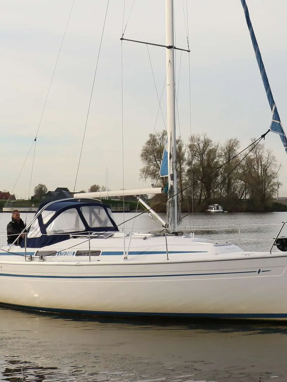Een witte zeilboot met blauwe strepen en een blauwe buiskap vaart op rustig water, met een persoon aan boord. — Bavaria 31