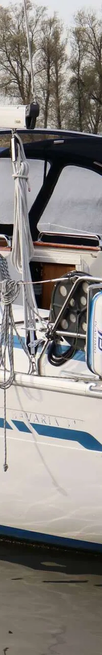 Een witte zeilboot met blauwe strepen en een blauwe buiskap vaart op rustig water, met een persoon aan het roer. — Bavaria 31
