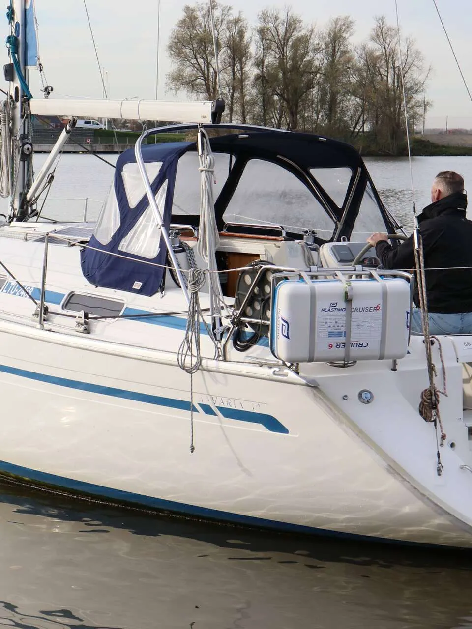 Een witte zeilboot met blauwe strepen en een blauwe buiskap vaart op rustig water, met een persoon aan het roer. — Bavaria 31