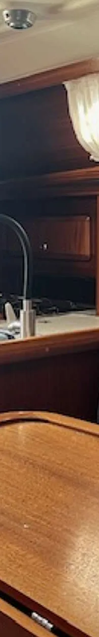 De foto toont het interieur van een boot met een houten tafel, een bank met grijze kussens en een kombuis met spoelbak.