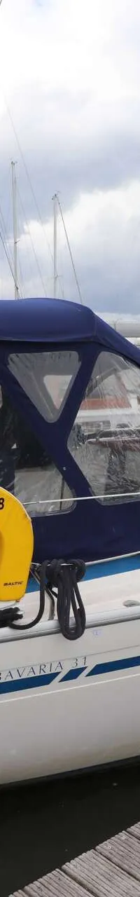 Een witte zeilboot met blauwe strepen en een blauwe buiskap ligt afgemeerd aan een houten steiger. — Bavaria 31
