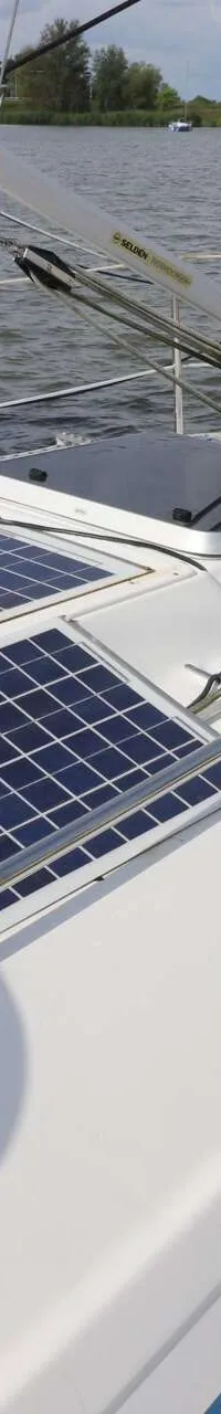 De foto toont het dek van een witte zeilboot met twee zonnepanelen en een luik, met op de achtergrond water en een andere…