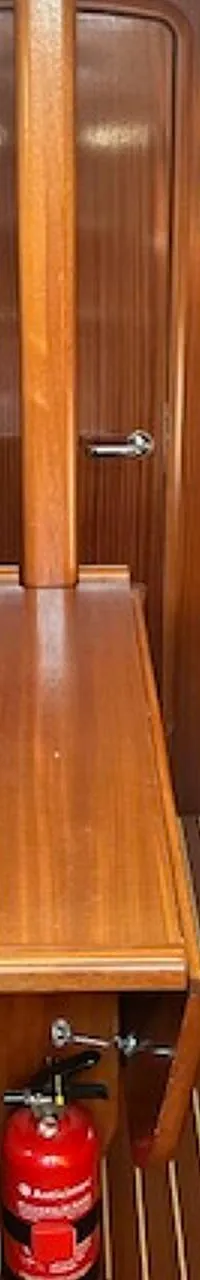 De foto toont het interieur van een kajuit met houten wanden, een tafel, twee banken met grijze kussens en een rode…