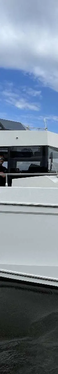 Een witte motorboot met een zwarte bimini en een persoon aan het roer vaart op water met een beboste oever op de achtergrond.