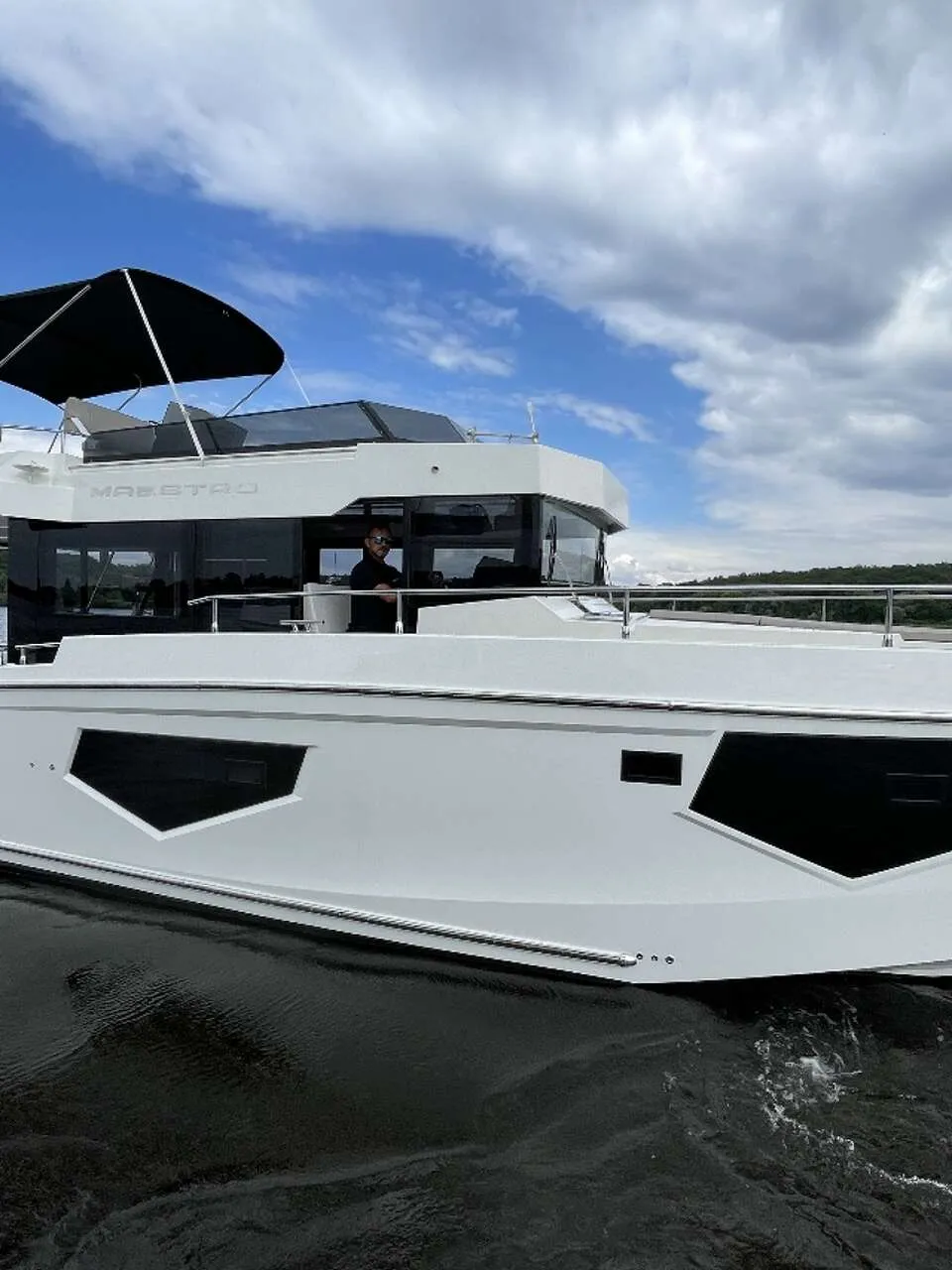 Een witte motorboot met een zwarte bimini en een persoon aan het roer vaart op water met een beboste oever op de achtergrond.