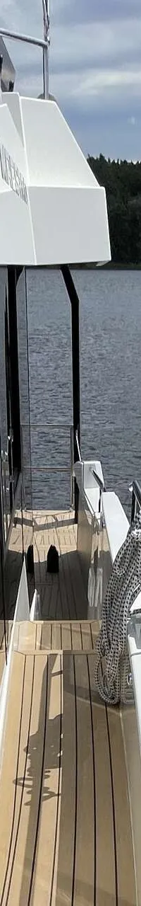 Een witte boot met een houten dek en donkere ramen vaart op een meer met beboste oevers onder een bewolkte lucht.