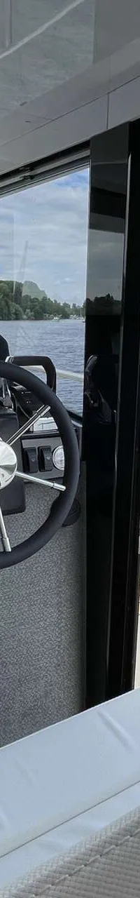 Een stuurhut met een stuurwiel, navigatiescherm en bedieningspanelen, met uitzicht op water en een beboste oever.