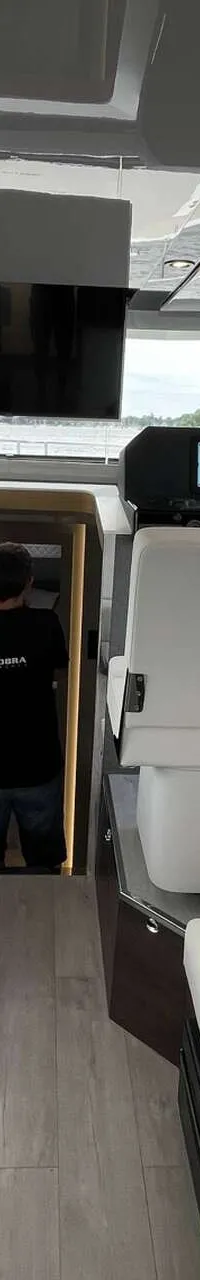 Een interieur van een boot met witte banken, een houten tafel, een stuurstand en een man in een deuropening.