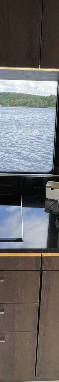Een bootkeuken met een spoelbak, kraan, kookplaat en kasten, met uitzicht op water en een beboste oever.