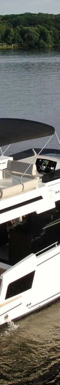 Een witte motorboot met een zwart dak en een houten achterdek vaart op een meer met bosrijke oevers.