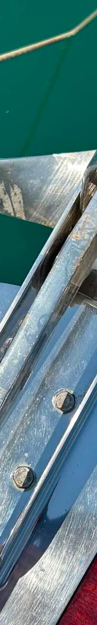 Een close-up van een glimmende, metalen ankerrol op een houten boeg, met groen water en een touw op de achtergrond.