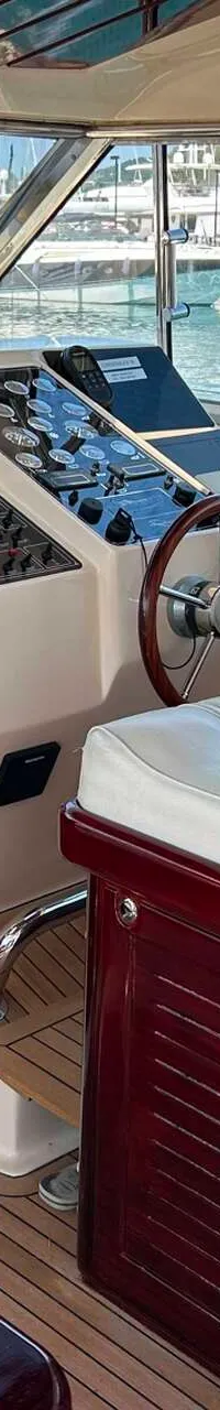 Een interieur van een boot met een stuurwiel, instrumentenpaneel, witte kussens en houten afwerking, met een jachthaven op…