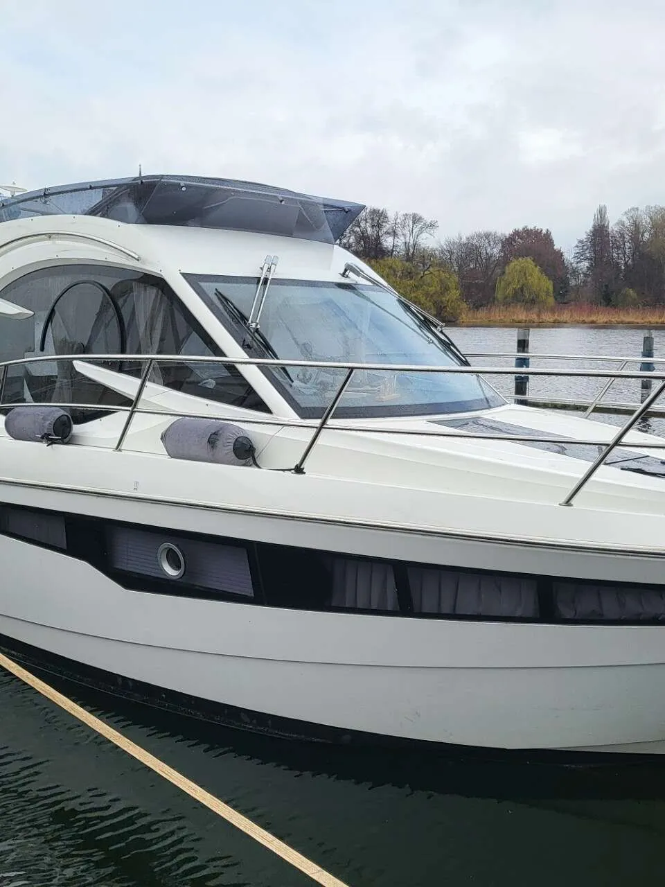 Een witte motorboot met een donkerblauwe kap en ramen, afgemeerd aan een steiger met een waterig landschap op de achtergrond.